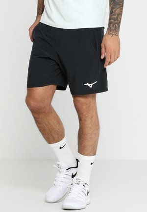 Mężczyzna ubrany w czarne sportowe szorty z białym logo, białe skarpetki i białe buty Nike, stojący na jednolitym tle.