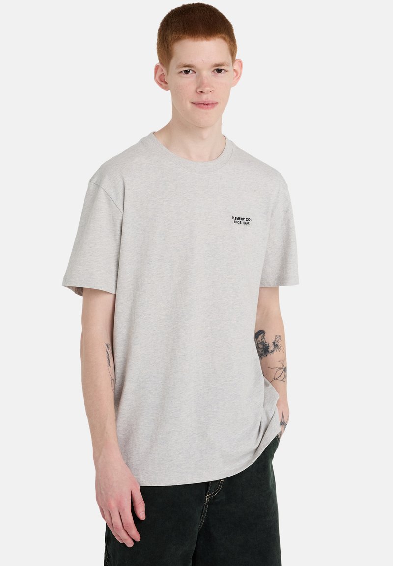 T-shirt gris clair à manches courtes avec un col rond et un petit logo noir sur la poitrine. Le tissu semble doux, avec une coupe décontractée.