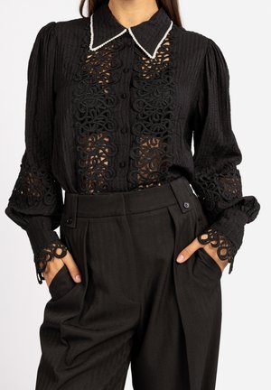 Blouse noire avec des découpes en dentelle sur le devant, les manches et les poignets, dotée d'un col bordé de blanc, associée à un pantalon noir taille haute avec les mains dans les poches.