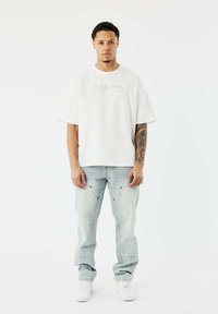 Camiseta blanca de corte XL con diseño bordado, combinada con jeans azul claro y zapatillas blancas, mostrando un ajuste relajado y un estilo casual.