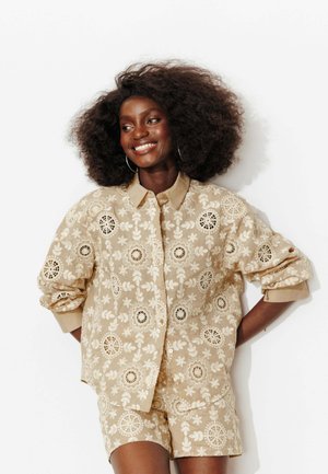 Femme avec coiffure afro naturelle souriante, portant une chemise beige en dentelle florale boutonnée et un short assorti, debout les mains derrière le dos.