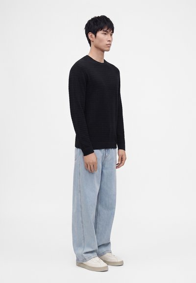 Un homme se tient face à gauche, portant un pull noir à manches longues, un jean bleu clair à jambes larges, et des baskets blanches sur fond blanc.