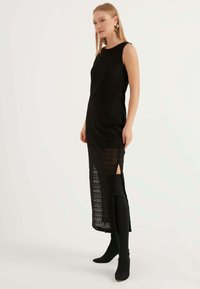 Abito nero in maglia con motivo a zigzag, senza maniche, con spacchi laterali e una silhouette lunga. Abbinato a stivali neri al ginocchio.