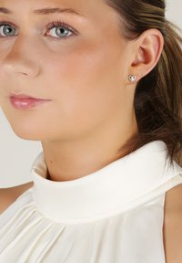 Hjärtförda silverörhängen med klara rhinestones, bärs av en person med ljus hy och ljusbrunt hår, klädd i en vit plisserad topp.