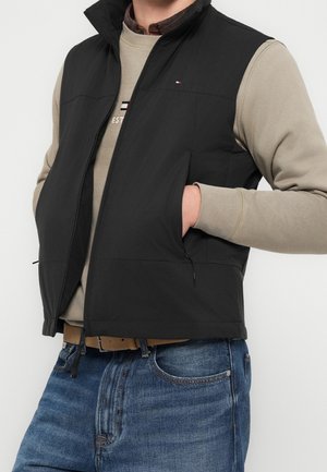 Homme portant un gilet noir sans manches zippé sur un sweat-shirt beige et un jean bleu, les mains dans les poches du gilet sur fond blanc.