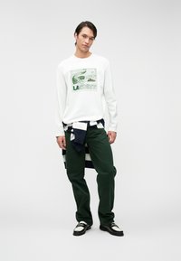 La camicia bianca a maniche lunghe presenta un grafico del logo Lacoste verde, abbinata a pantaloni verde scuro e a un maglione a righe blu navy e bianche legato in vita.