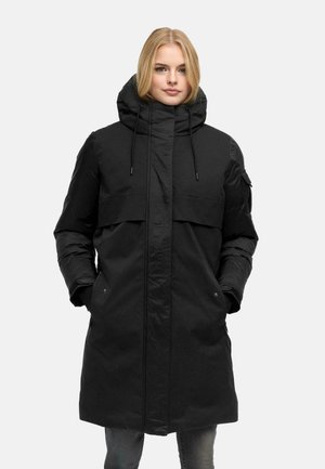 Femme portant un long manteau matelassé noir avec un col montant et une capuche, les mains dans les poches, debout devant un fond clair uni.