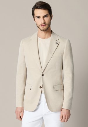 Mężczyzna w beżowej marynarce na jasnej koszuli i białych spodniach, stojący na tle jednolitego beżowego tła, patrzący przed siebie.