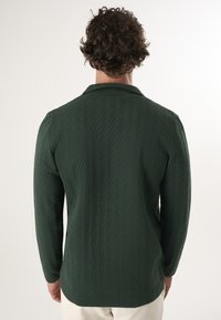 Maglietta a maglia verde scuro con un motivo a diamante testurizzato, maniche lunghe e colletto. Presenta una vestibilità rilassata e dettagli con fessure laterali.