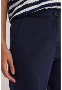 Mână odihnindu-se lângă pantaloni bleumarin croiți, cu închidere cu nasture, purtați cu un top tricotat alb cu dungi bleumarin.