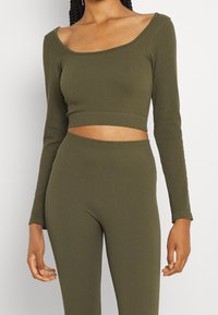 Top corto a maniche lunghe a coste di colore verde oliva abbinato a leggings a vita alta coordinati. Presenta un ampio scollo e una silhouette aderente.