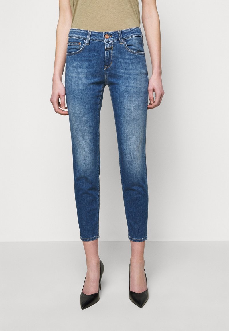 Woman wearing slim-fit, ankle-length blue jeans with faded details and black pointed high heels, standing against a plain background.