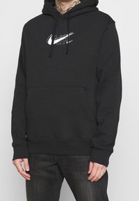 Černá mikina s kapucí vyrobená z bavlněné směsi, obsahující přední klokankovou kapsu a bílé logo Nike. Šňůrky na kapuci.