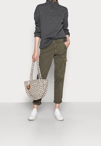 Grijze gewatteerde pullover met hoge kraag, olijfgroene cargo-broek met zakken, en een patroon geweven tas. Witte sneakers maken de look af.