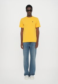 Tricou galben din bumbac, cu mâneci scurte, având un logo mic brodat, combinat cu blugi de culoare bleu deschis, cu croială largă și adidași albi.