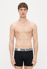 Czarne bokserki z białym elastycznym paskiem w talii z napisem „ONLY&SONS”. Tkanina wydaje się gładka i dopasowana. Brak innych szczegółów.