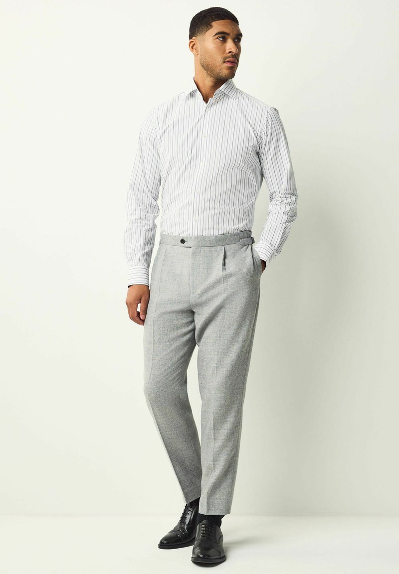 Camisa de vestir a rayas blancas y grises combinada con pantalones gris oscuro a medida que presentan un bolsillo tipo hendidura y un detalle de un solo botón. Zapatos de vestir negros.