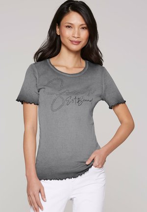 Woman wearing a gray short-sleeve shirt with black ruffled edges and black script text, paired with white pants, smiling at camera.