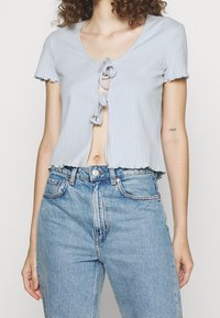 Žena v světle modrém krátkém rukávovém topu s vázáním vpředu a světle modrých džínech s vysokým pasem, ukazující břišní část a tetování na ruce.