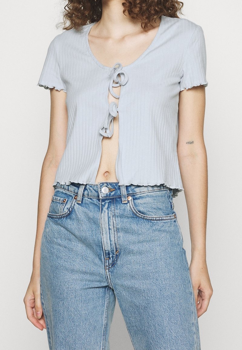 Žena v světle modrém krátkém rukávovém topu s vázáním vpředu a světle modrých džínech s vysokým pasem, ukazující břišní část a tetování na ruce.