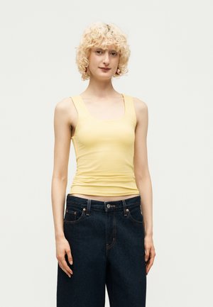 Jeune personne aux cheveux blonds bouclés, portant un débardeur jaune et un jean large bleu foncé, debout devant un fond uni.