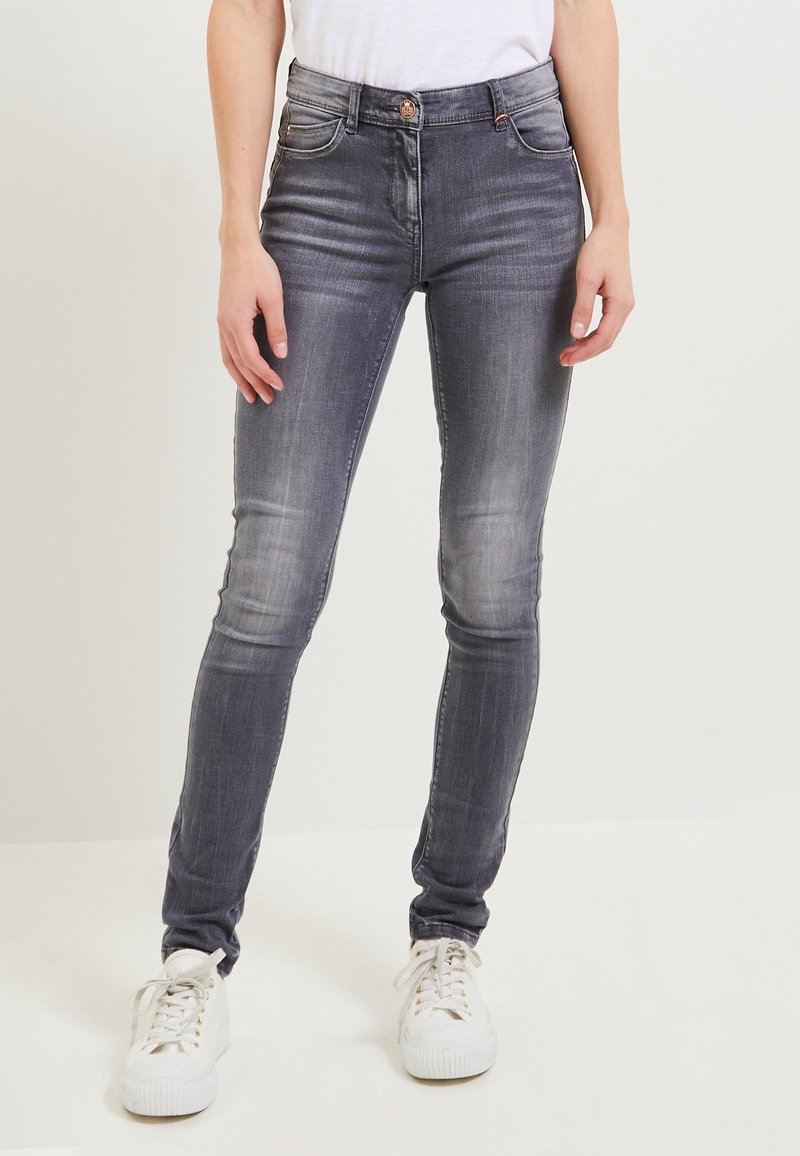 Jean gris délavé slim porté avec des baskets blanches et un haut blanc, debout devant un fond clair uni.