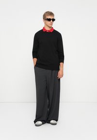Young man wearing black sweater over red polo, gray wide-leg trousers, white sneakers, and black sunglasses, standing with hands in pockets.