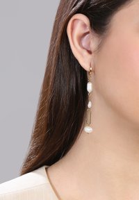 Goldfarbene Tropfenohrringe mit weißen Perlen, einem ovalen Link und einer leicht unregelmäßigen Form, die von einem kleinen Ring befestigt sind.