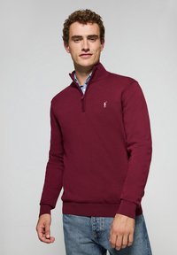 Suéter burdeos de cuello con cremallera y puños acanalados, con un pequeño logo en el pecho. Llevado sobre una camisa de cuadros.