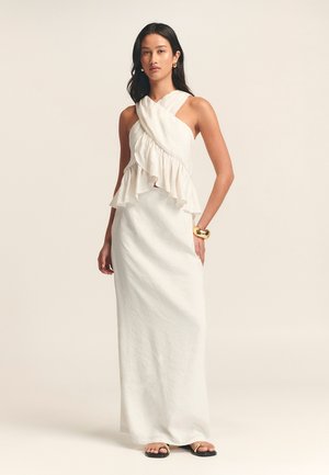 Abito maxi bianco con orlo arricciato e scollo incrociato. Il tessuto sembra leggero e con una texture particolare. Sandali con dettagli dorati completano il look.
