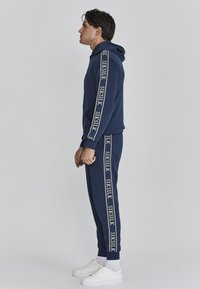 Giovane in tuta con cappuccio blu navy con il testo "SIKSILK" sulle maniche e strisce sui pantaloni, indossando calzini bianchi e sneaker bianche, in piedi di lato.