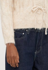 Μπεζ πουλόβερ πλεκτό με σχέδιο καλωδίου, ζωνάκι με κορδόνι και υφή, σε συνδυασμό με σκούρο τζιν παντελόνι με αντιθετική ραφή.
