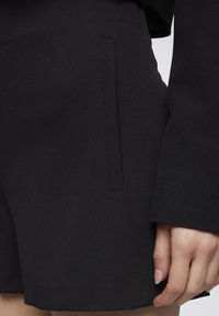Pantaloni corti neri dal design sartoriale, con una tasca laterale e una superficie in tessuto liscia. La mano si appoggia delicatamente sul bordo del capo.