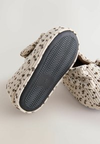 De petites chaussures pour bébé en beige doux avec des pois bruns, l'une montrant une semelle noire texturée, toutes deux ornées d'un nœud en tissu sur le dessus.