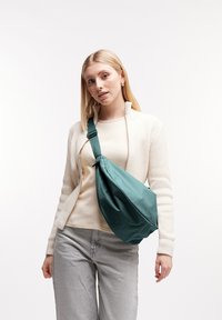 Teal tygslingväska med knutdetalj och justerbar rem, bärs över en ljus tröja och kombineras med grå jeans, mot en enfärgad bakgrund.