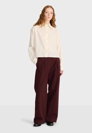 Jeune femme aux longs cheveux roux bouclés portant une chemise crème boutonnée, un pantalon marron foncé à jambes larges et des chaussures noires sur un fond uni.