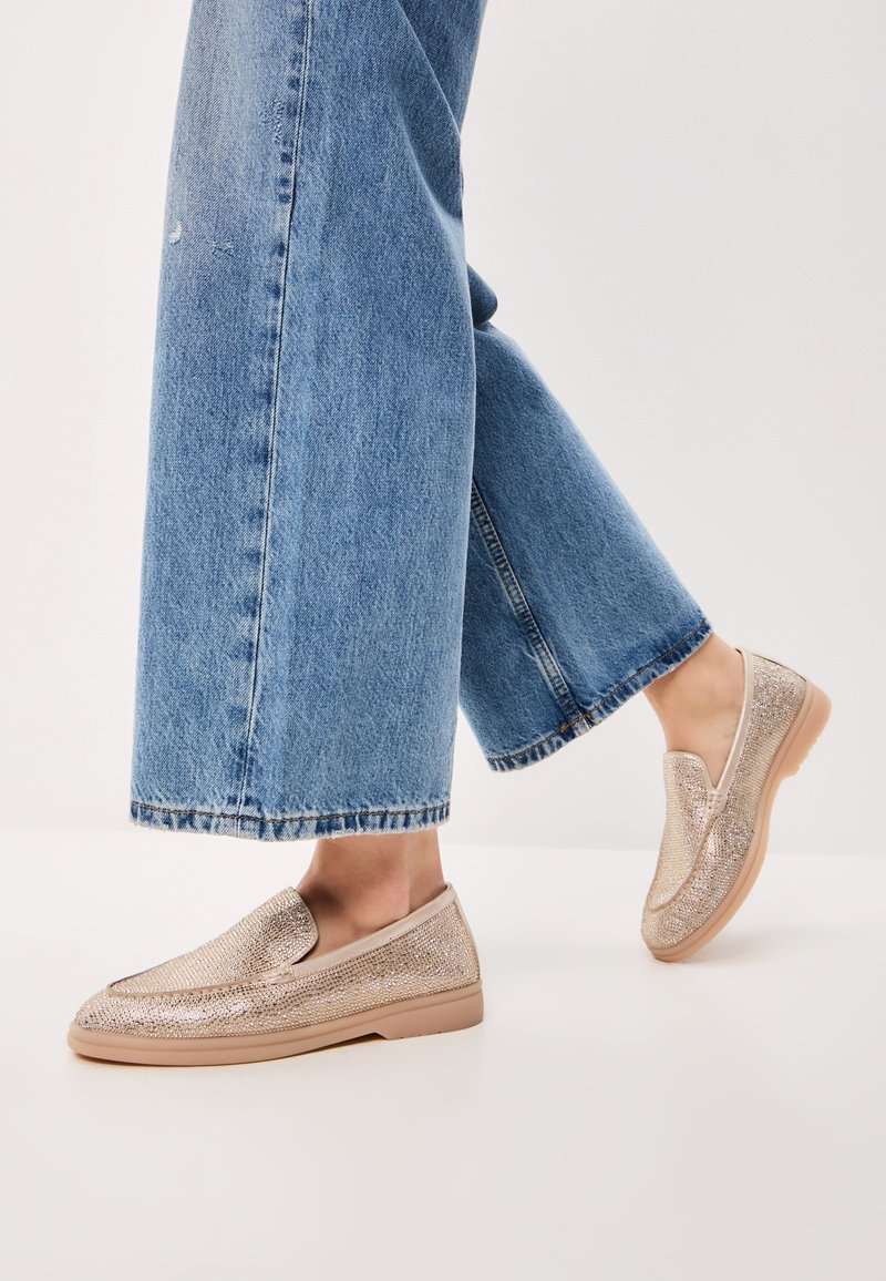 Metalické zlaté slip-on boty s hladkým povrchem, zaoblenou špičkou a gumovou podrážkou, nošené s volnými modrými džíny.