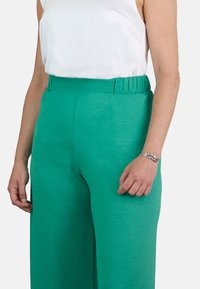 Pantalones anchos de pierna amplia en verde, ligeros y con cinturilla elástica. Tela texturizada visible; combinados con una sencilla blusa sin mangas blanca y una pulsera de plata.