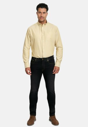 Camisa de boton en beige de manga larga, con un bolsillo en el pecho, combinada con unos jeans negros ajustados y unos zapatos de cordones marrones. Diseño simple y clásico.