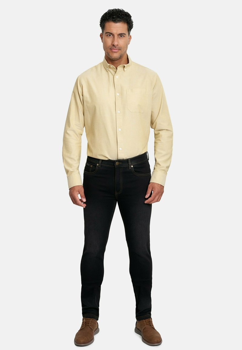 Camisa de boton en beige de manga larga, con un bolsillo en el pecho, combinada con unos jeans negros ajustados y unos zapatos de cordones marrones. Diseño simple y clásico.