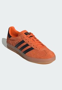 Adidași din suede portocalie cu dungi negre, vârf rotund, toc texturat negru și talpă din cauciuc semi-transparentă. Închidere cu șireturi și detalii cu logo vizibile.