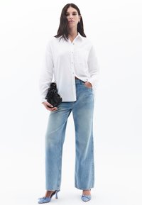 Weißes Hemd mit Knopfverschluss, hellblaue weit geschnittene Jeans und blaue Absatzschuhe. Schwarze Handtasche mit gerafften Details.