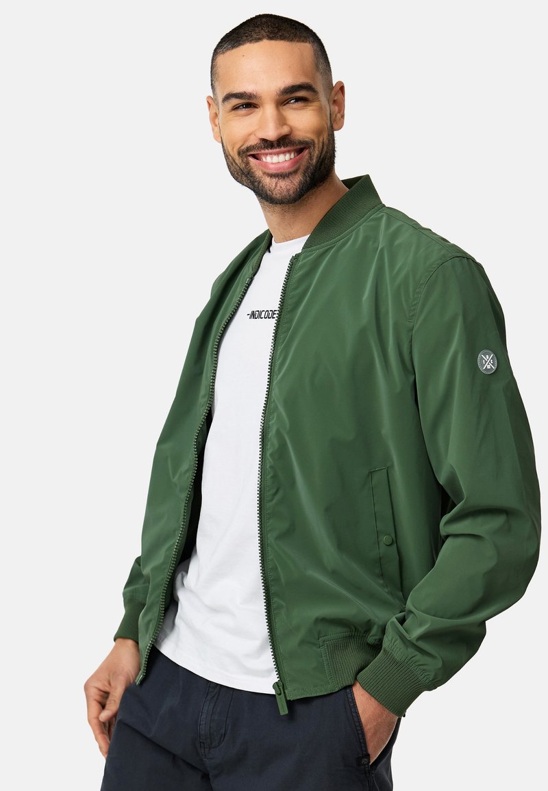 Grüne Bomberjacke mit glatter Textur, gerippten Bündchen und Reißverschluss. Hat einen Logopatch auf dem linken Ärmel. Trägt man über einem weißen Hemd.