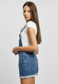 Denim overalls with adjustable straps, light blue wash, rolled hem, and metal button accents. Paired with a fitted white t-shirt.