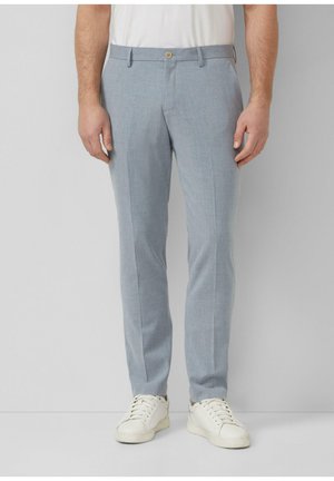 Mężczyzna ubrany w jasnoniebieskie, dopasowane spodnie i białe trampki, stojący na tle jednolitego, jasnoszarego tła.