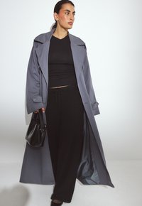 Moteris, vilkinti ilgą pilką tranšėjinį paltą ant juodos palaidinės ir kelnių, laiko juodą rankinę, stovinti prieš baltą fono.
