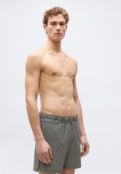 Jeune homme aux cheveux bouclés portant un short à rayures verticales noires et blanches, debout devant un fond gris clair uni.