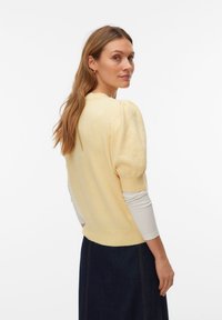 Femme aux longs cheveux portant un pull jaune clair à manches courtes bouffantes sur une chemise blanche à manches longues et une jupe foncée, tournée légèrement vers l'arrière.