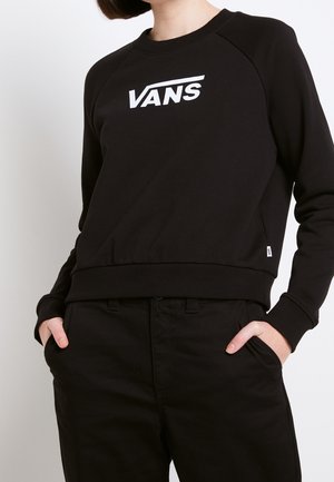 Persona con sudadera negra de Vans con logo blanco, pantalones negros, con las manos en los bolsillos, vista desde el cuello hasta las caderas sobre fondo blanco.