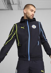 Giacca nera con cappuccio, con accenti blu e verde neon, apertura con zip e logo del Manchester City sul petto. Texture liscia.